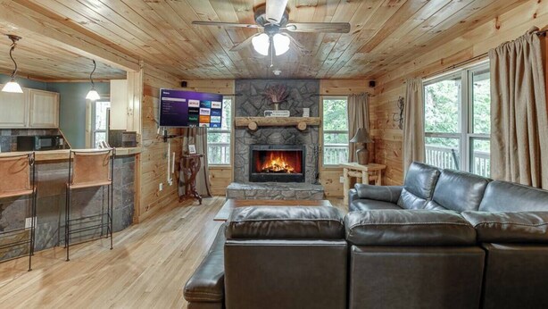 A cabin interior with a stone fireplace, wooden ceiling, a leather sofa, a bar area, and a television.