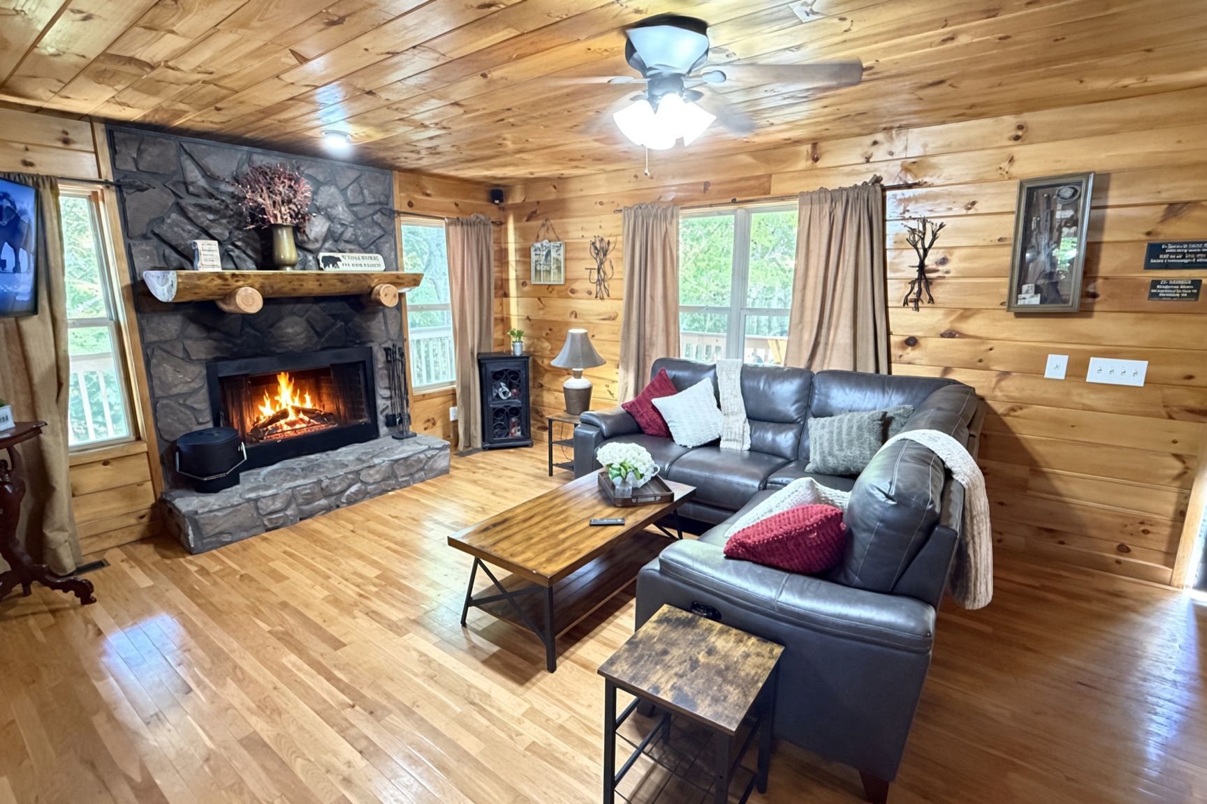 A cozy cabin interior with a stone fireplace, leather sofa, wooden flooring, and a ceiling fan.