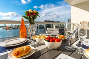 A dining table with plates, bowls of fruit, and wine glasses, overlooking a cityscape and water. - Spacious condo on high floor with amazing bay & city views. WIFI + Parking (Sunny Isles Beach)