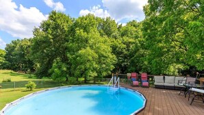 An outdoor swimming pool with a wooden deck, surrounded by trees and a fenced area.