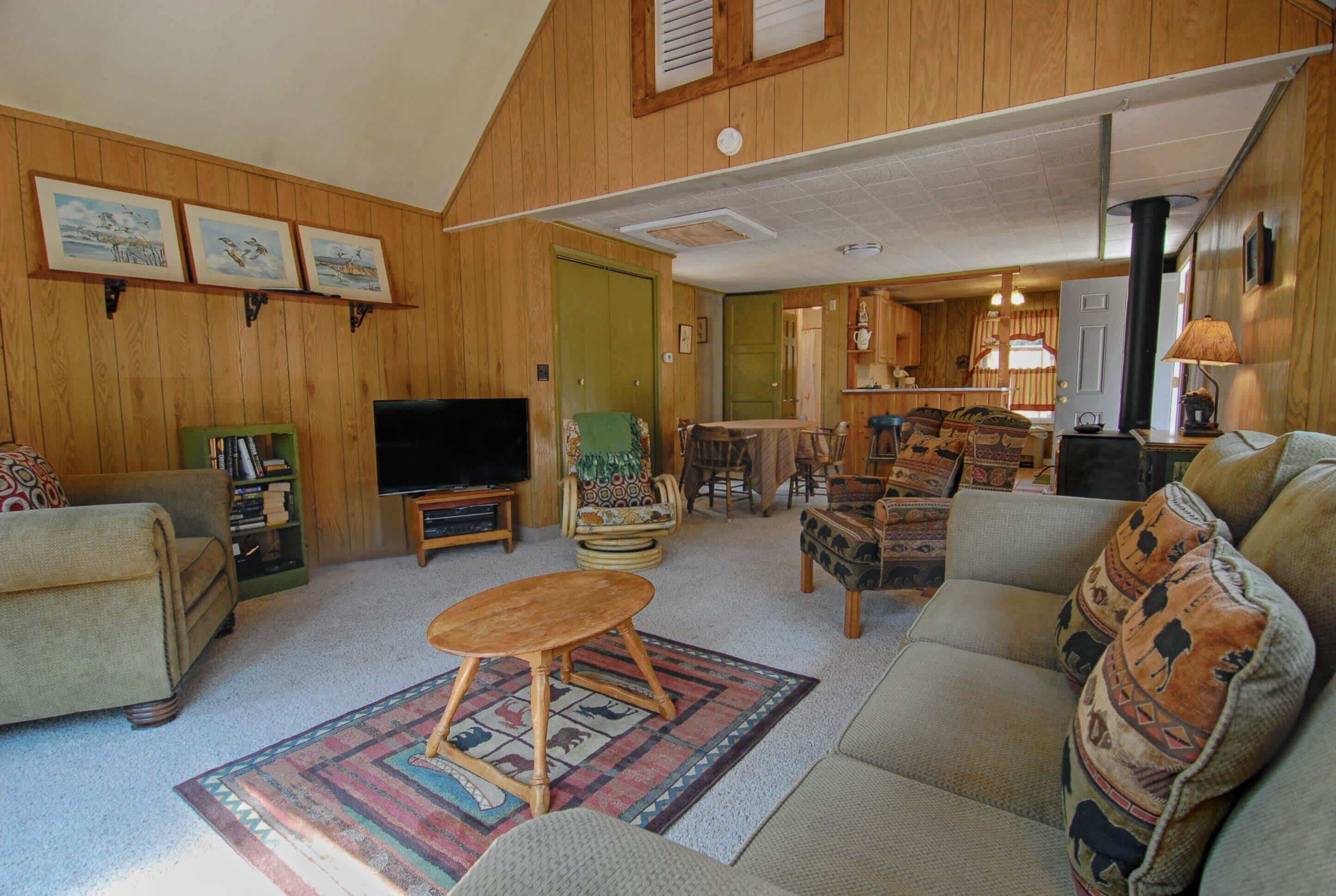 A wooden-paneled living room with a sofa, armchair, television, and a dining area with chairs and a table.