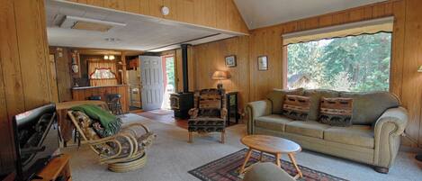 A cozy living room with a sofa, armchair, and television, wooden walls, and a large window.