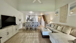 Un vaste espace de vie comprenant un téléviseur à écran plat, un ventilateur de plafond, une table à manger avec des chaises, un canapé et un escalier menant à un autre niveau.