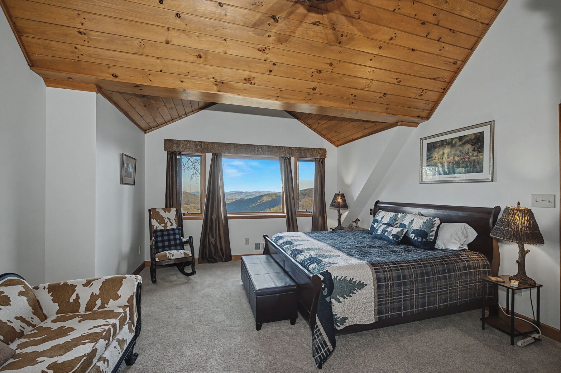 A bedroom with a large bed, a chair, a bench, a table, and a window with a view of the mountains.