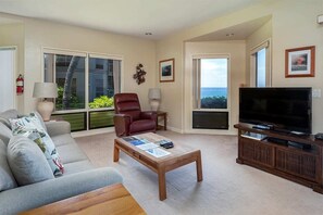 A living room with a sofa, armchair, coffee table, TV, and a view through a sliding door. - Ocean’s Edge w/Sunset View Lanai! WiFi, Modern Kitchen, TV, Washer/Dryer Kanaloa 2001 (KAILUA-KONA)
