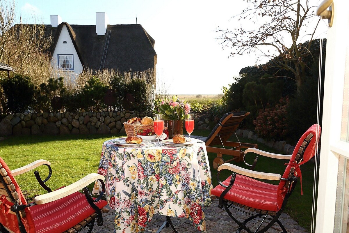An outdoor dining area with a table set for two, surrounded by red chairs, a thatched-roof house in the background, and a stone wall.