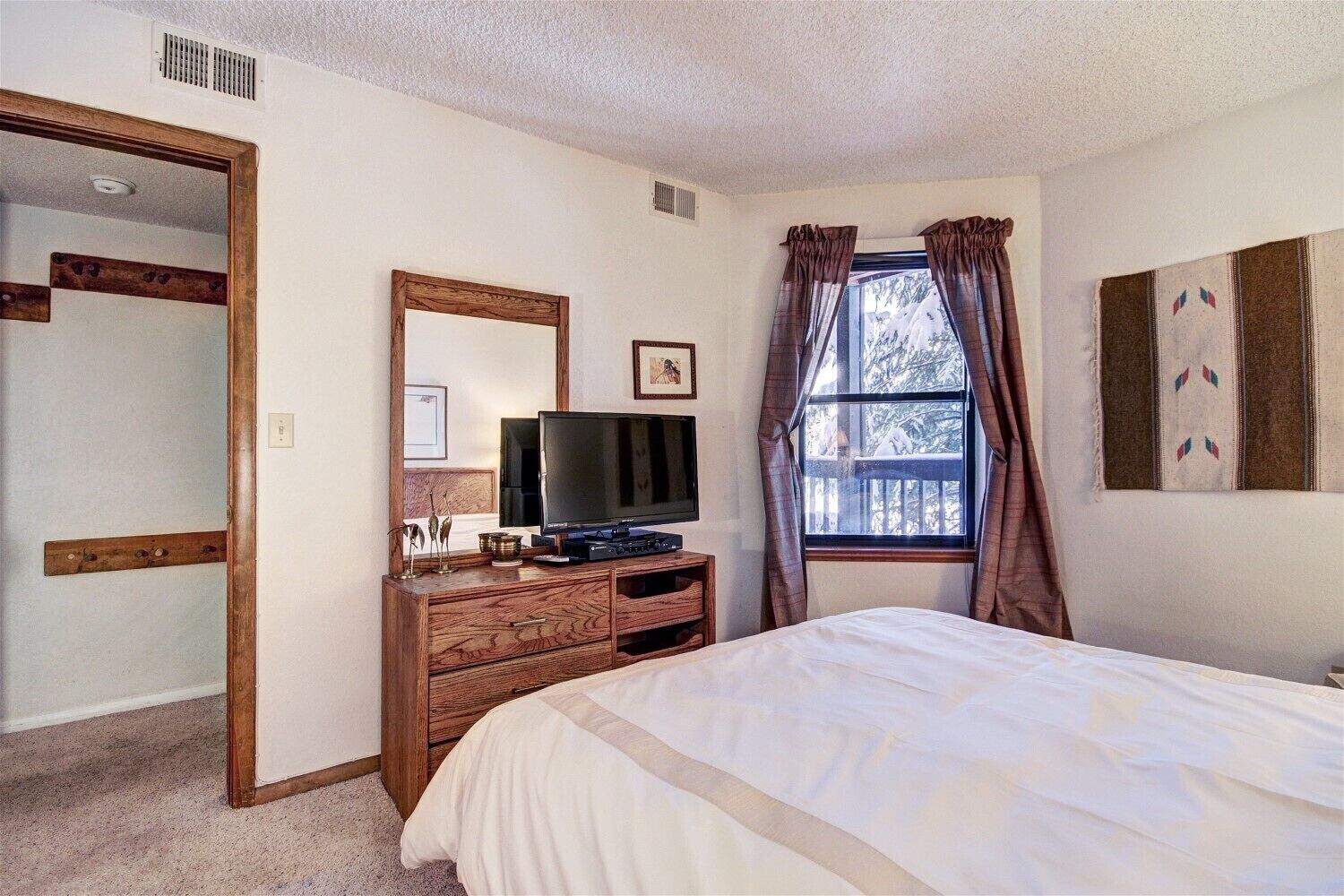 A bedroom with a bed, a TV on a wooden dresser, a window with curtains, and a mirror on the wall.