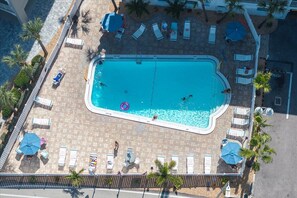 An aerial view of a rectangular swimming pool with lounge chairs and umbrellas, surrounded by palm trees and a tiled deck.