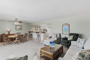 A living room with a dining area, a kitchen in the background, and a view of the outdoors through a window.