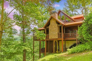 A cabin with a stone chimney, wooden deck, and a view of the forest.