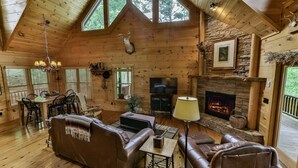 A log cabin interior with a fireplace, leather sofas, a television, and a dining area.