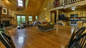 A wooden cabin interior with a dining table, chairs, a fireplace, a kitchen area, and a staircase leading to an upper level.