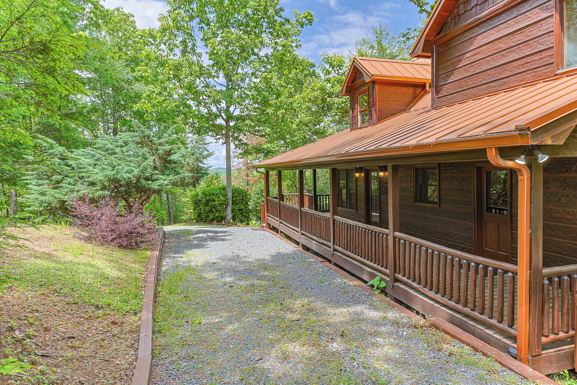 A wooden cabin with a porch, surrounded by trees and a gravel path.