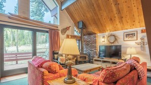 A cozy living room with a fireplace, a television, and a large window overlooking a forested area. - Chalet #6, Charming Cabin Perfect Location steps t (Mammoth Lakes)