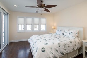 Quarto com uma cama grande, um ventilador de teto e três janelas com persianas.