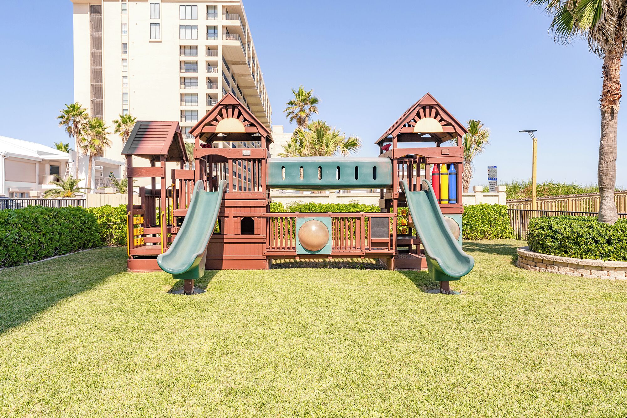 Un parque infantil con dos toboganes y una estructura para escalar, rodeado de césped y palmeras, con un edificio alto al fondo.