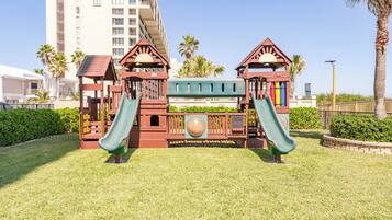 Un parque infantil con dos toboganes y una estructura para escalar, rodeado de césped y palmeras, con un edificio alto al fondo.