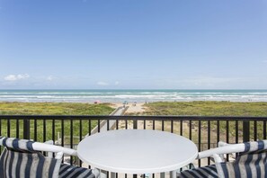 En balkong med ett vitt bord och randiga kuddar, med utsikt över en strand där folk sitter och det finns parasoller.