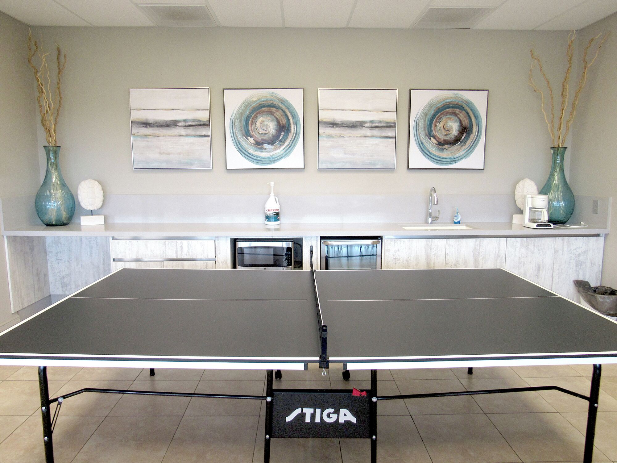 A ping pong table with a TIGA logo, a kitchen area with a sink, and three abstract paintings on the wall.