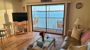A living room with a television, a coffee table with a vase, a sofa, and a view of the sea through large windows.