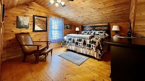 Um quarto aconchegante estilo cabana, com teto de madeira, cama com colcha estampada, uma cadeira, uma escrivaninha e um ventilador de teto.