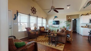 Ein Wohnzimmer mit Deckenventilator, Fernseher, Rattanmöbel und einer Küchenzeile.