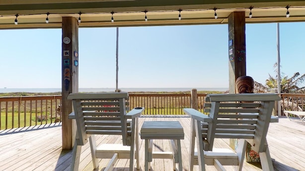 Eine Holzterrasse mit zwei weißen Stühlen und einem kleinen Tisch mit Blick auf einen Strand.