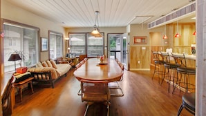 A spacious dining area with a large wooden table surrounded by chairs, a kitchen area with bar stools, and a cozy seating area with a sofa and armchairs. - Idle Hours ~ Fantastic River Views & Walk to Downtown! Classic Russian River! (Guerneville)