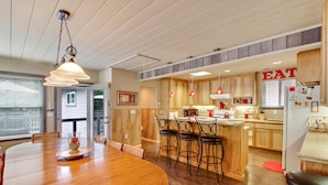A modern kitchen with wooden cabinets, a dining area with a large wooden table, and a kitchen island with bar stools. - Idle Hours ~ Fantastic River Views & Walk to Downtown! Classic Russian River! (Guerneville)