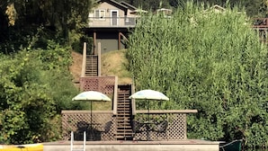 A house with a deck overlooking a lake, surrounded by dense greenery. - Idle Hours ~ Fantastic River Views & Walk to Downtown! Classic Russian River! (Guerneville)