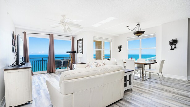 Un salon spacieux avec un grand canapé blanc, un coin repas avec une table et des chaises, et un balcon offrant une vue sur l’océan.