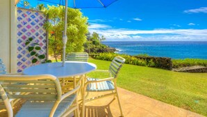 Eine Terrasse mit Tisch und Stühlen unter einem blauen Sonnenschirm, mit Blick auf einen Strand mit klarem, blauem Wasser und einer felsigen Küstenlinie.