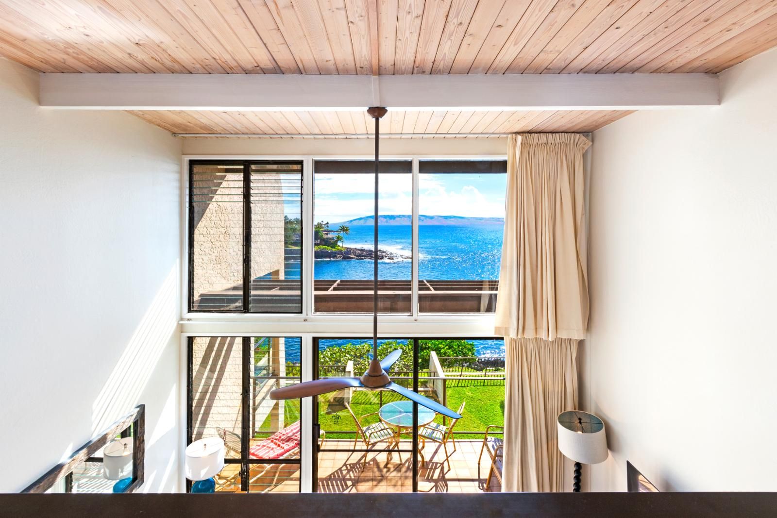A room with a large window overlooking a scenic ocean view, a wooden ceiling, and a balcony with outdoor furniture.