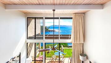 A room with a large window overlooking a scenic ocean view, a wooden ceiling, and a balcony with outdoor furniture.