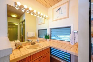 Un baño con tocador, un espejo grande con iluminación, una ventana con persianas y un cuadro en la pared.
