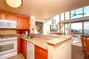 A kitchen with granite countertops, wooden cabinets, a microwave, stove, and a sink. There is a large window with a view of the ocean. - Napili Point | Oceanfront 2 BR | Water Front | Car Incl. w/ 6+ Nights | NAP-C18 by KBM (Lahaina)