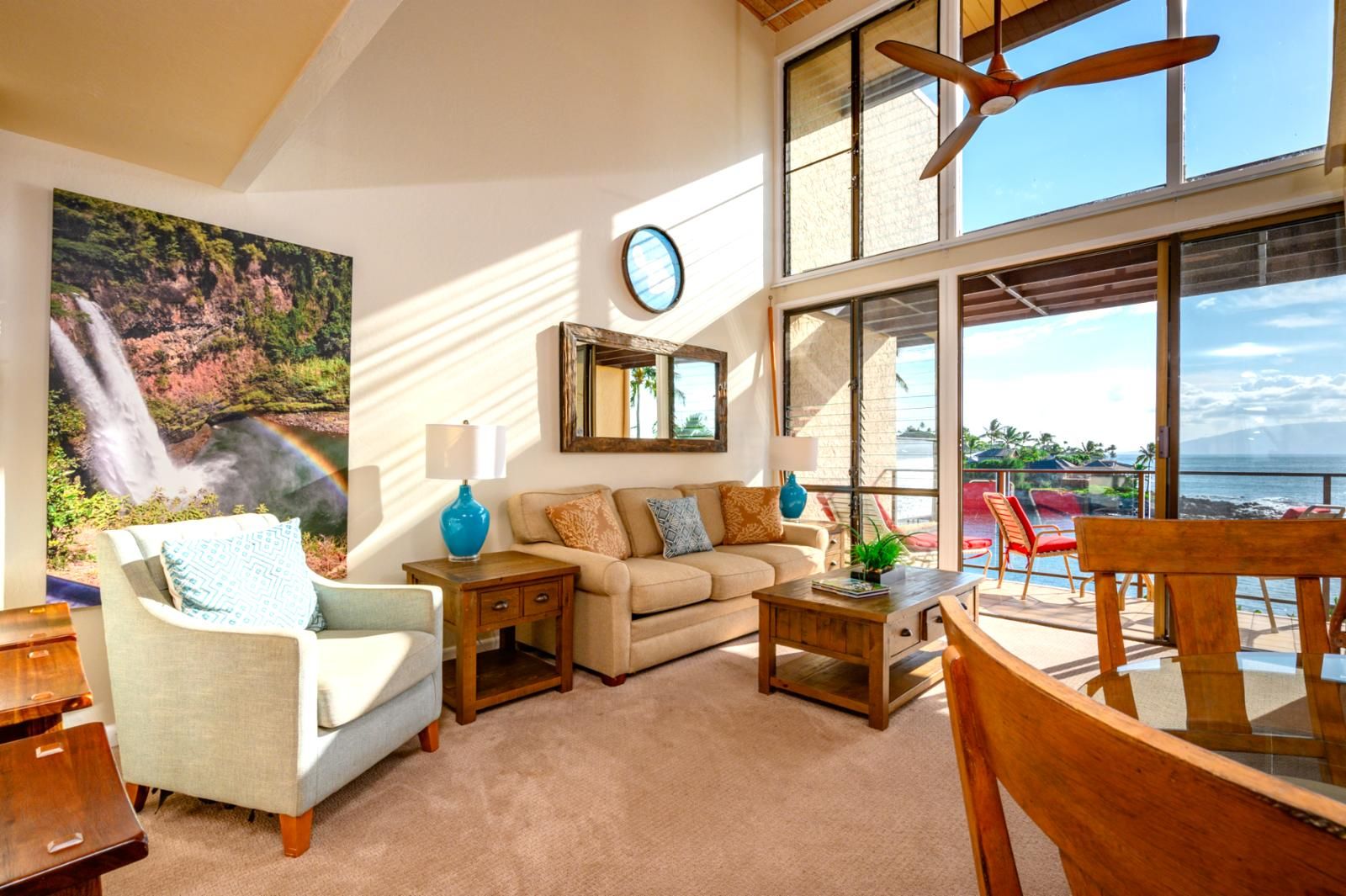 A living room with a sofa, coffee table, and chairs, a large window with a view of the ocean, and a painting of a waterfall.