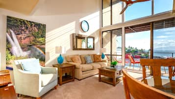 A living room with a sofa, coffee table, and chairs, a large window with a view of the ocean, and a painting of a waterfall.