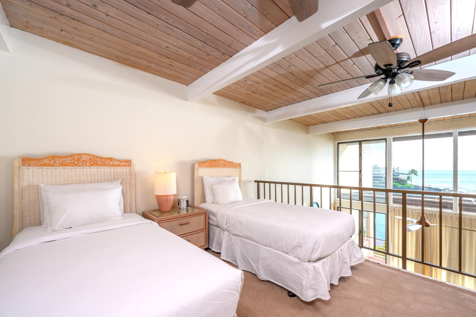 Habitació d'hotel amb dos llits, un ventilador de sostre, una tauleta de nit amb una làmpada i un balcó amb vista a l'oceà.
