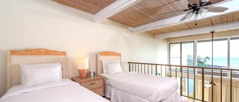 A hotel room with two beds, a ceiling fan, a nightstand with a lamp, and a balcony with a view of the ocean.