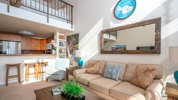 A spacious living area with a high ceiling, a kitchen with wooden cabinets, a large mirror, and a comfortable sofa with cushions.