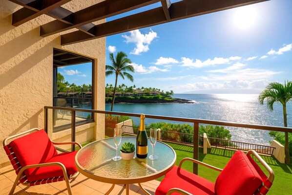 A balcony with a glass table, red chairs, and a view of the ocean and a tropical landscape.