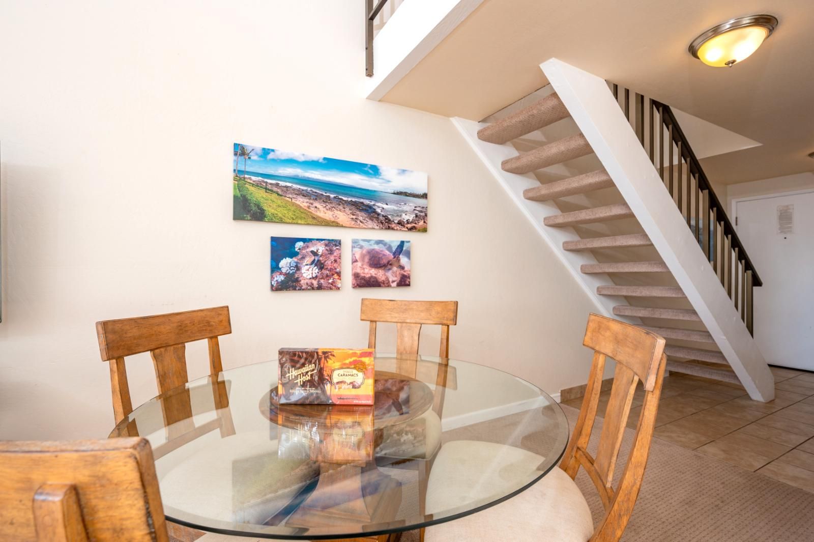 A dining area with a glass table and wooden chairs, a staircase with a white railing, and three framed pictures on the wall.