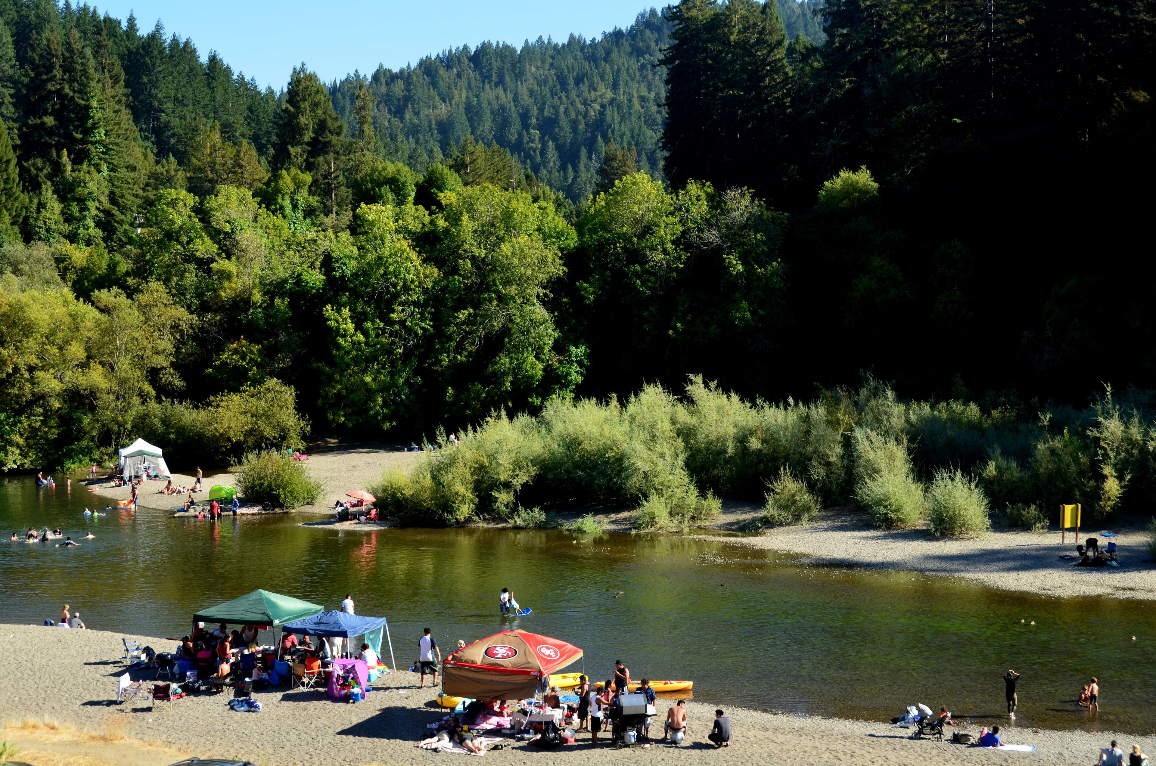 Camping junto al río con tiendas de campaña, zona de barbacoa y personas disfrutando de actividades acuáticas.