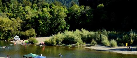 Camping junto al río con tiendas de campaña, zona de barbacoa y personas disfrutando de actividades acuáticas.