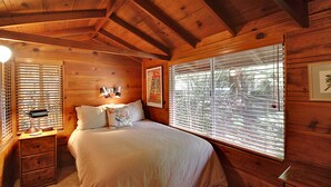 Une chambre dans une cabane en bois, avec un lit, une table de chevet et une fenêtre équipée de stores.