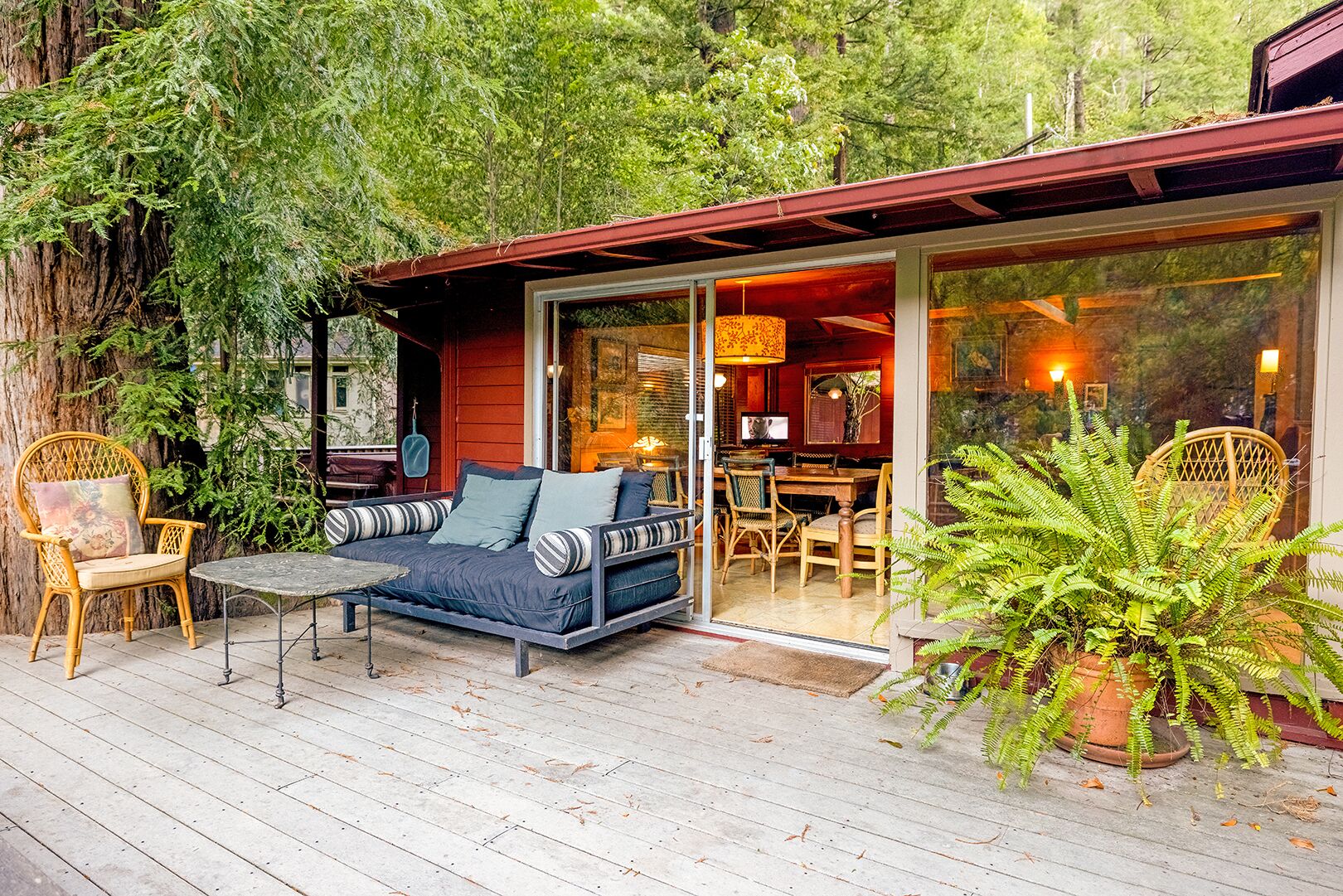 Un rincón exterior acogedor con sofá, sillas y una mesa, rodeado de abundante vegetación y una terraza de madera.