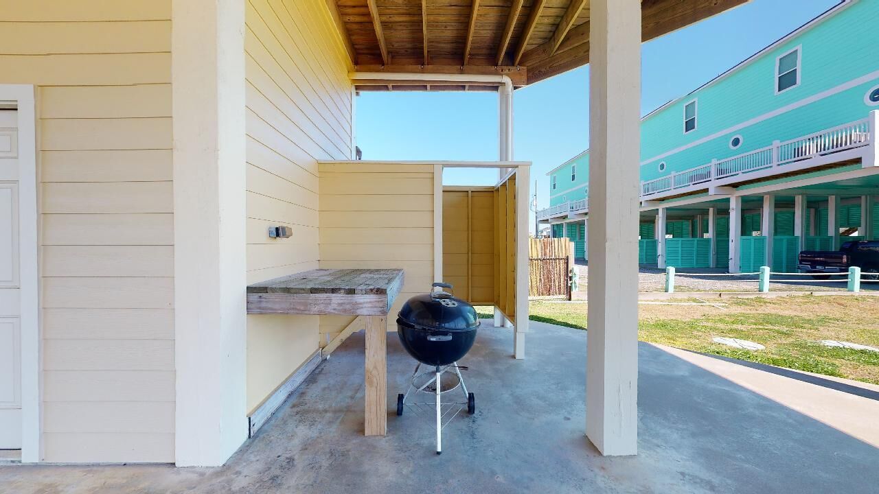 Ein überdachter Außenbereich mit Grill, Holztisch und einem Gebäude im Hintergrund.