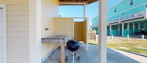 A covered outdoor area with a grill, a wooden table, and a building in the background.