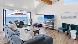 A modern living room with a large gray sofa, a teal armchair, a wooden coffee table, and a TV displaying a beach scene. - Beautiful UPPER LEVEL Pet-Friendly Oceanfront Duplex P1111-1 (Oceanside)